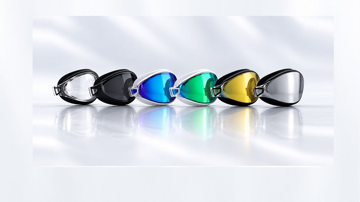 Six swim goggles arranged in a row the POD AquaLuxe Swim Goggles with black or white frames and reflective lenses in clear, black tint, blue, green, gold and silver, arranged in a row on a reflective white surface.