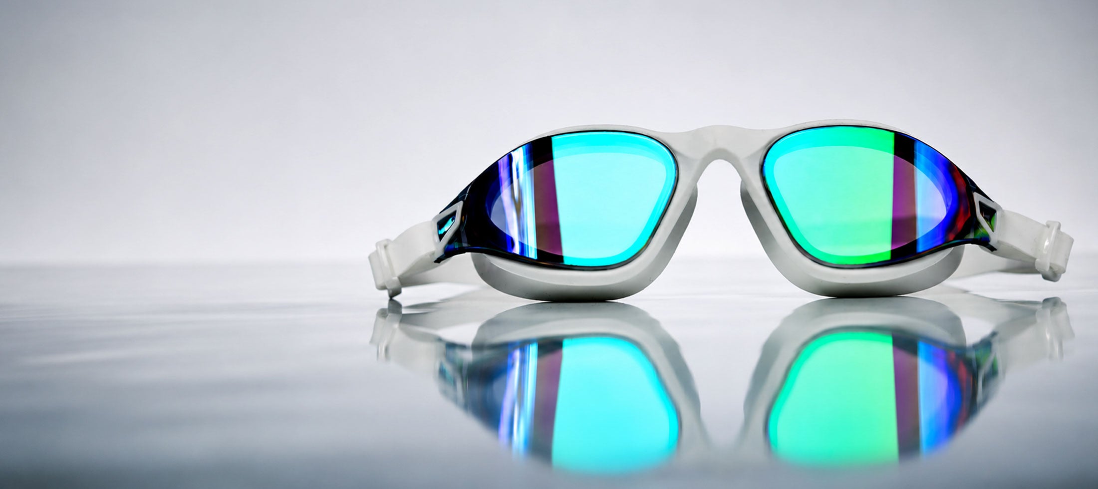 White Aqualuxe swim goggles with vibrant blue and green mirrored lenses, resting on a reflective grey surface against a light grey background.