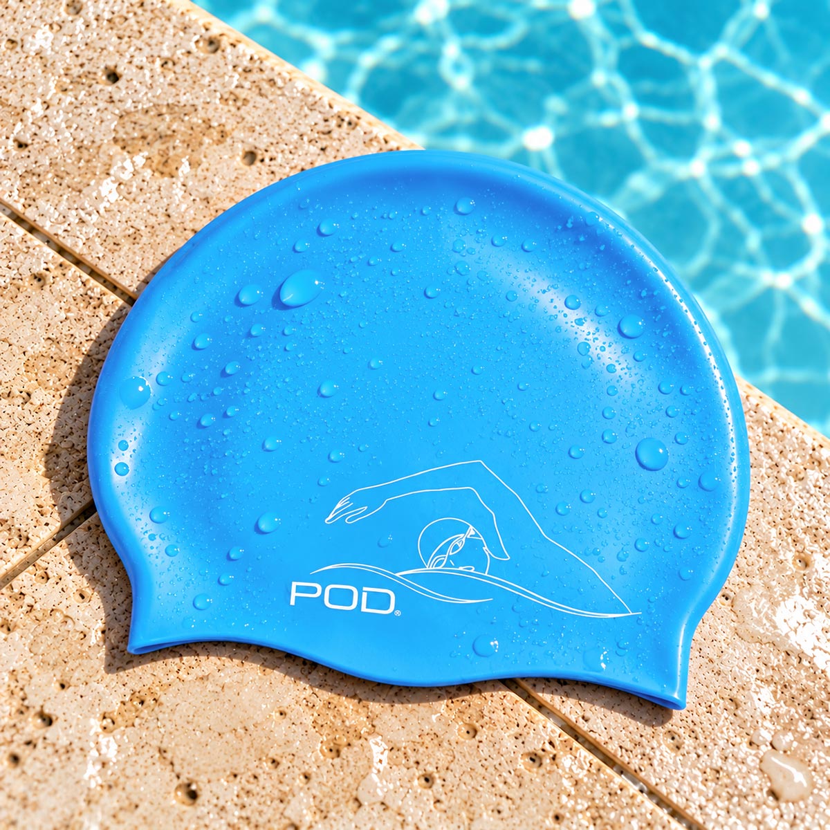 A bright blue silicone swim cap with water droplets, featuring a white line-art swim logo and the brand name 'POD.' resting on a beige stone pool deck with blue pool water in the background.