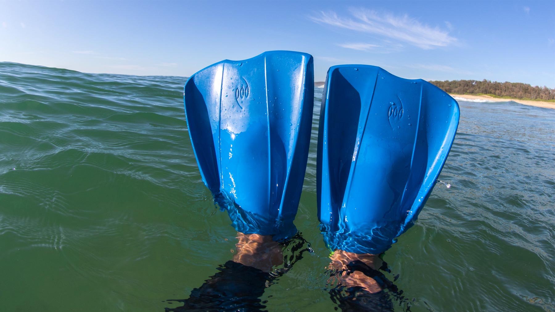 A rider's feet wearing blue POD PF2 swim fins raised above the water surface in the ocean, with a sandy beach and tree line visible in the background under a clear blue sky