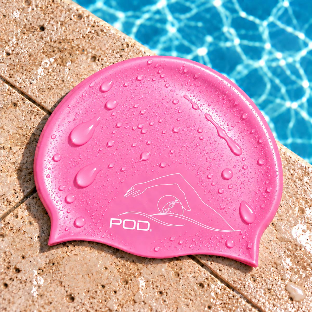 A pink silicone swim cap with water droplets, featuring a white swim graphic and the brand name 'POD.' on the front, resting on a pool deck with blue pool water in the background.