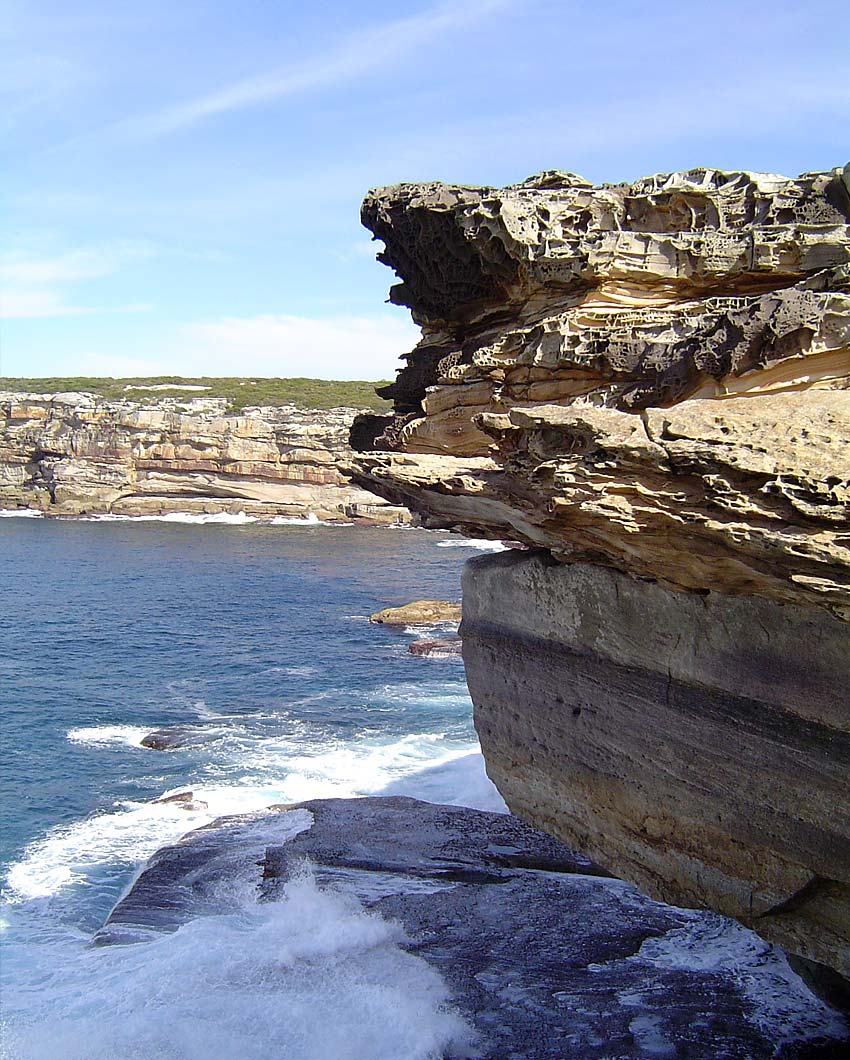 A coastal landscape photograph showing layered tan and brown rock cliffs over deep blue ocean water with white surf under a pale blue sky.