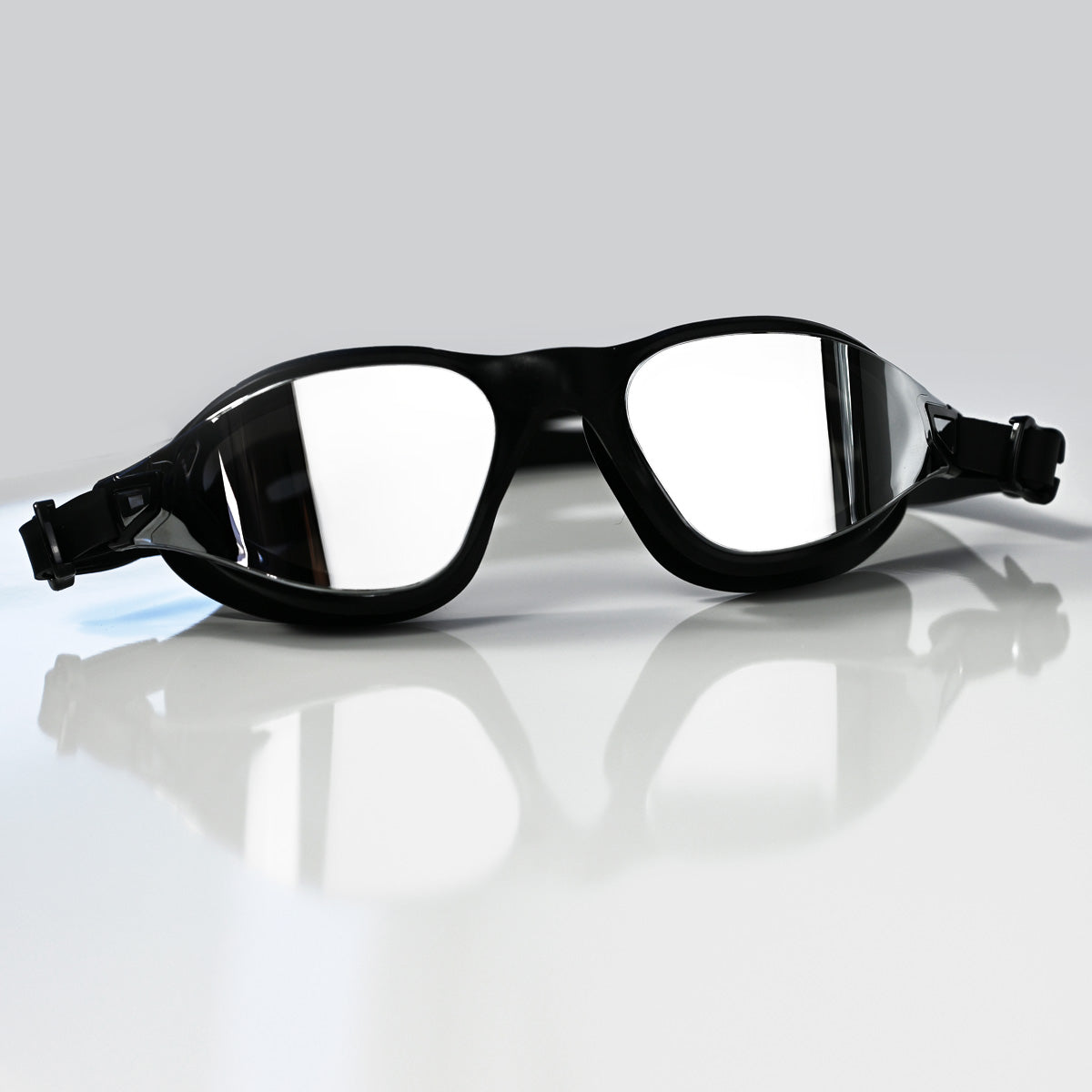Black swim goggles with silver mirror reflective lenses, resting on a glossy white surface with a soft reflection underneath.