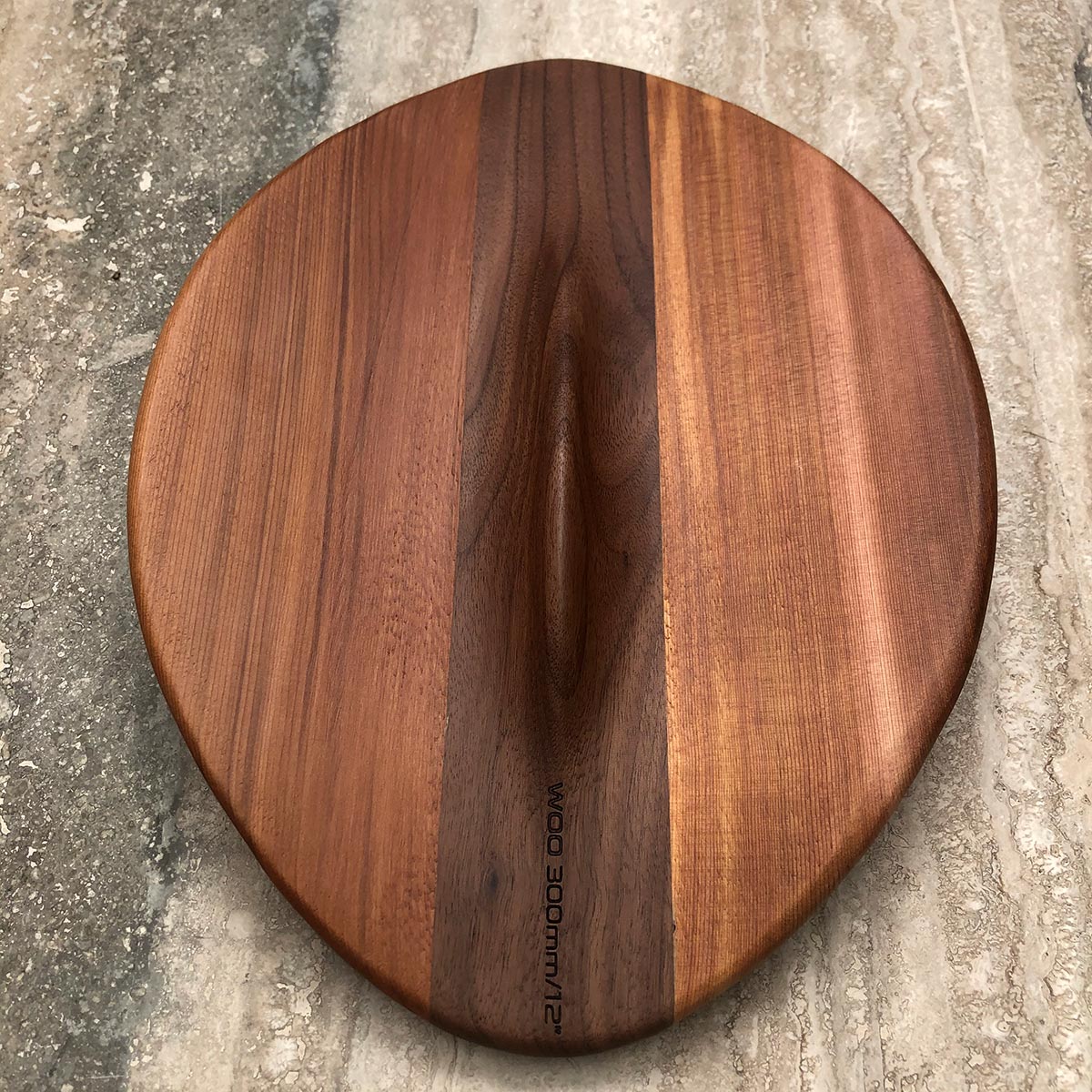 An organic-shaped wooden body surfing board with a natural wood grain pattern and a central handle, placed on a textured concrete surface. The brand name 'WOO 300mm x 2' is printed vertically near the bottom edge.