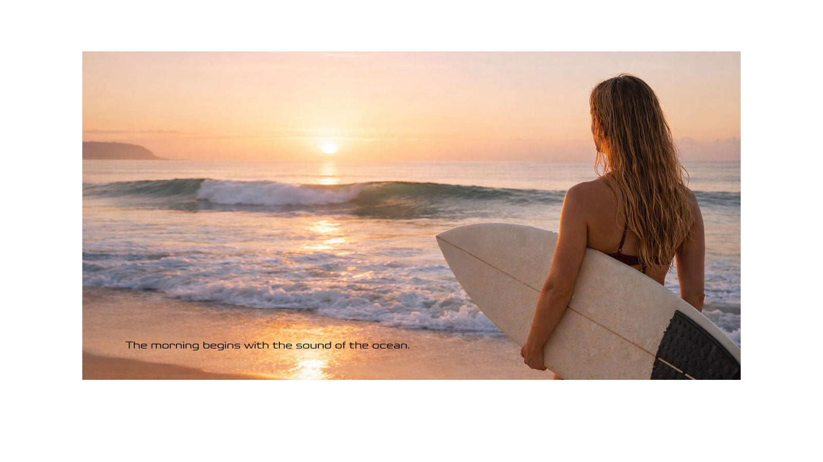  A surfer standing on the beach at sunrise holding a white surfboard, looking out at small waves breaking along the shore with a headland in the background