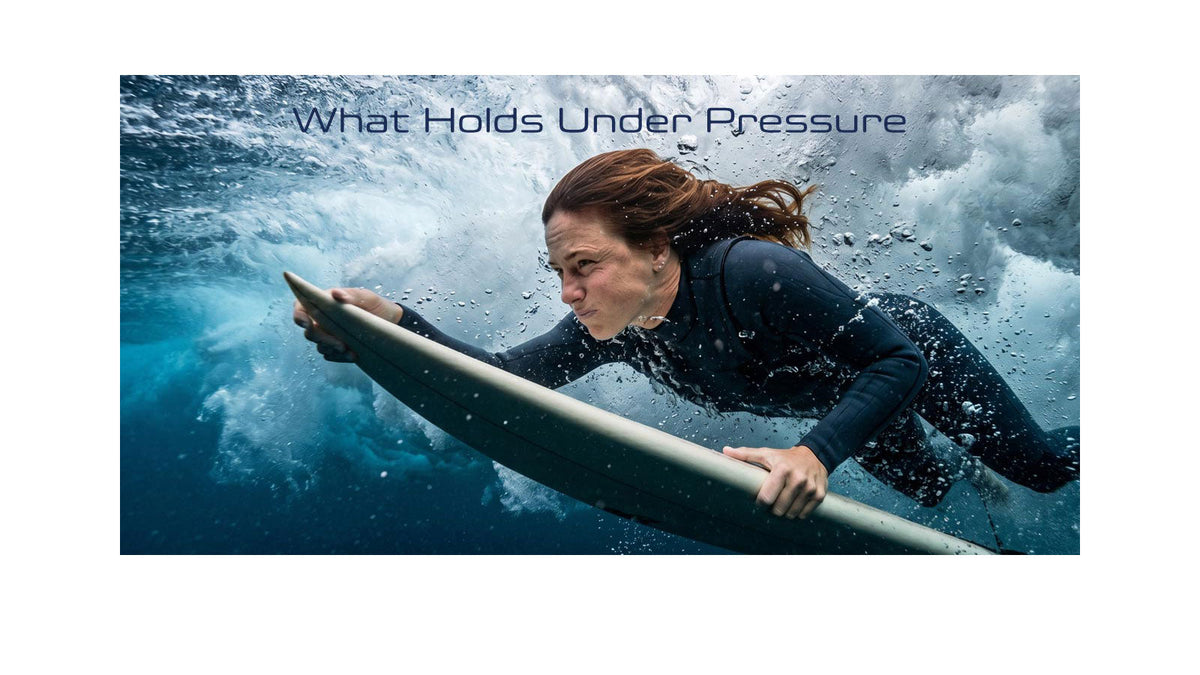 A female surfer duck-diving underwater through a breaking wave while holding a surfboard, wearing a black wetsuit and ear plugs, with the text "What Holds Under Pressure" overlaid on the image