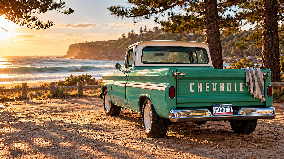 A vintage teal and cream Chevy pickup truck parked on a dirt beachside road with the ocean, trees, and sunset in the background. The driver-side door is open, and a striped towel hangs from the tailgate.