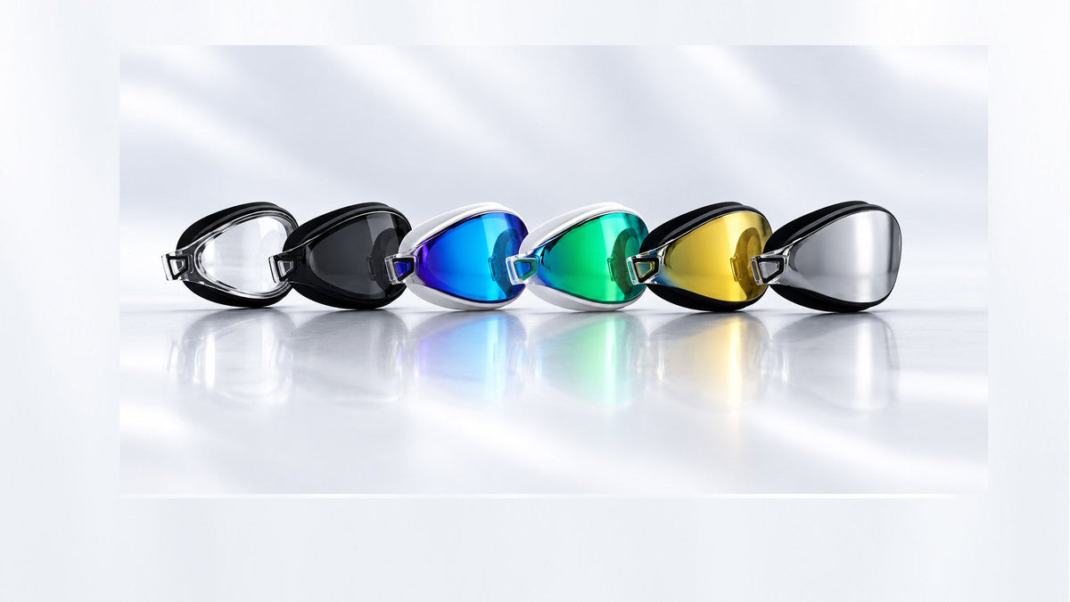 Six swim goggles arranged in a row POD-AquaLuxe Swim Goggles with black or white  frames and reflective lenses in clear, black tint, blue, green, yellow, and silver, arranged in a row on a reflective white surface.