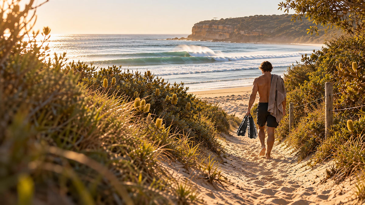 A man walks barefoot down a sandy path between green dunes toward the ocean at sunset, carrying a towel and POD PF2 Body Surfing Swim fins.