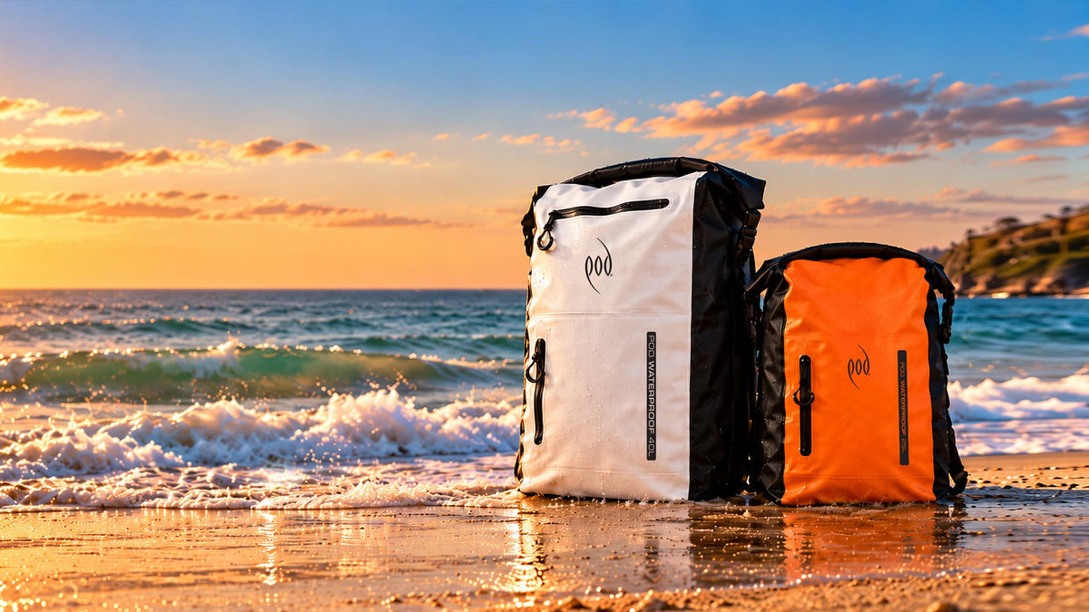 Two POD waterproof backpacks on wet sand at the water's edge at sunset, a white 40L and an orange 25L, with waves breaking behind them.