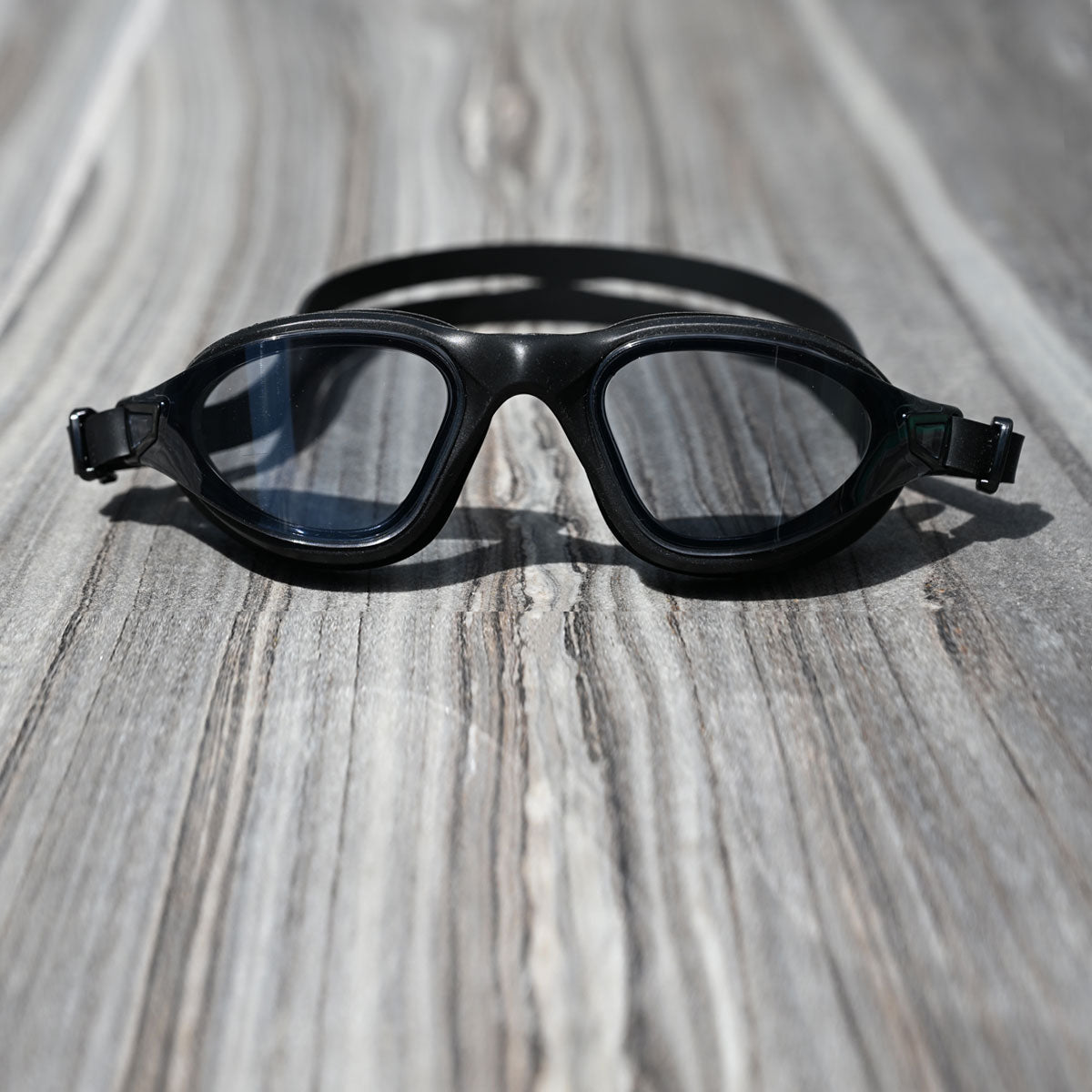 POD AquaLuxe swim goggles with black tinted lenses and black frame resting on a weathered timber surface in natural light