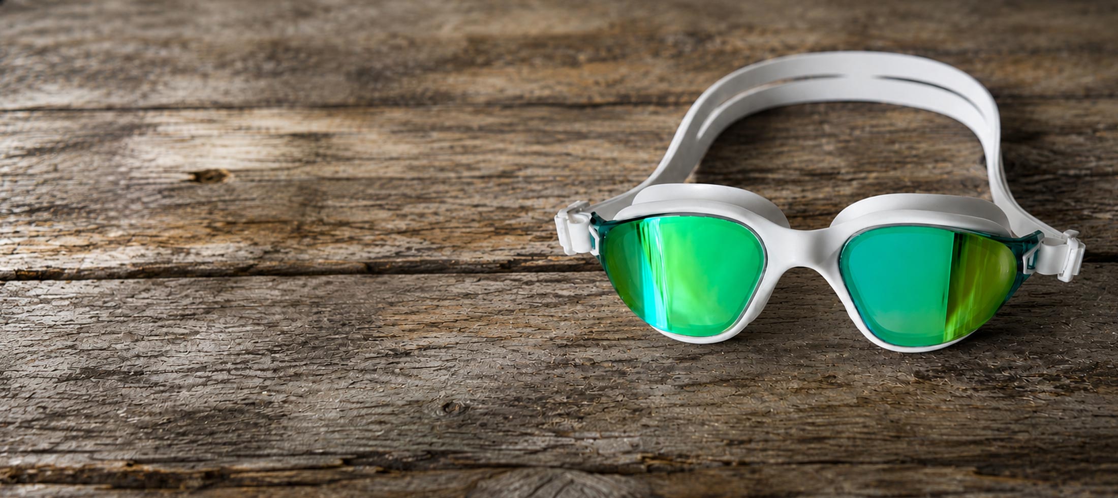 White swim goggles with green mirrored lenses resting on a rustic wooden surface.