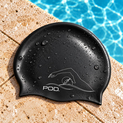 A black silicone swim cap with water droplets, featuring a white line art logo of a swimmer and the brand name 'POD' on the front, resting on a beige stone pool deck with blue pool water in the background.