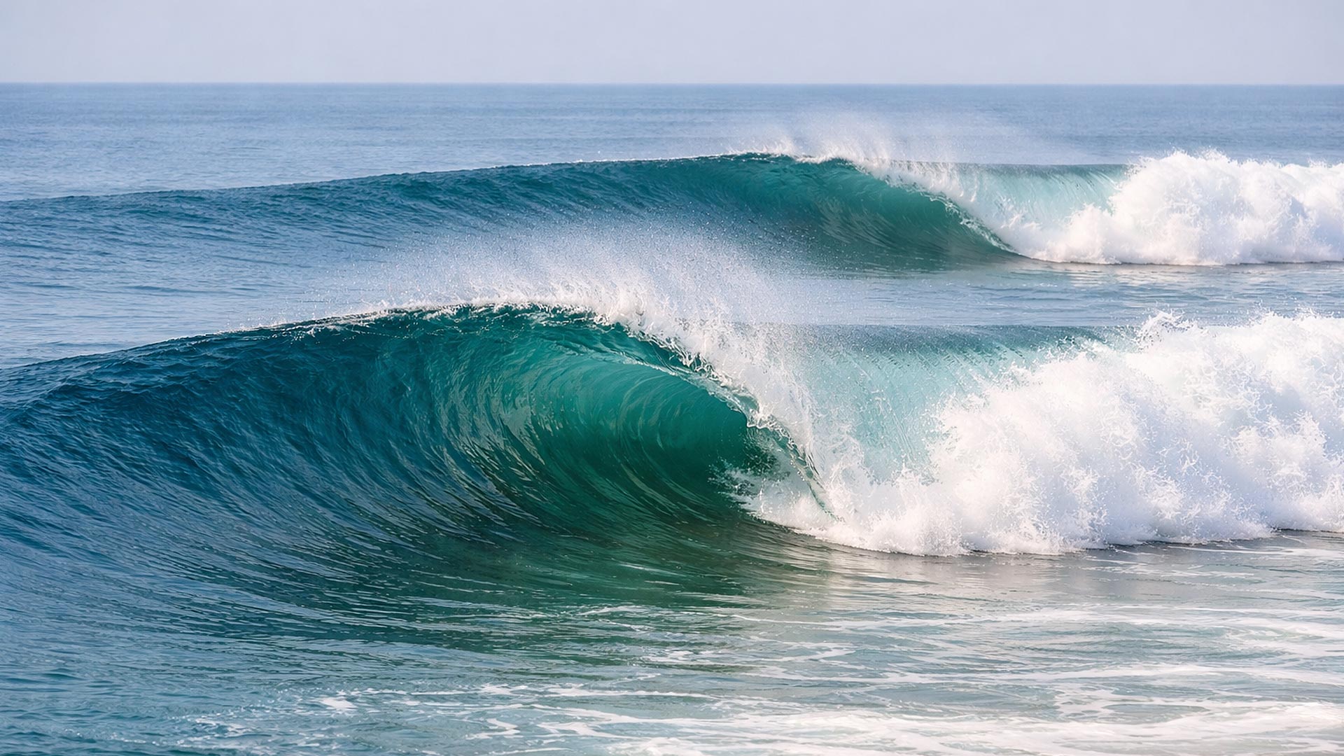 A large turquoise ocean wave curls and breaks with white foam under a pale blue sky.