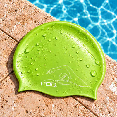A bright green silicone swim cap with water droplets, featuring a white POD logo, resting on a tan pool deck with blue pool water in the background.