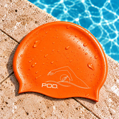 An orange silicone swim cap with water droplets, featuring a white line art logo and the brand name 'POD.' resting on a stone pool deck with blue pool water in the background.