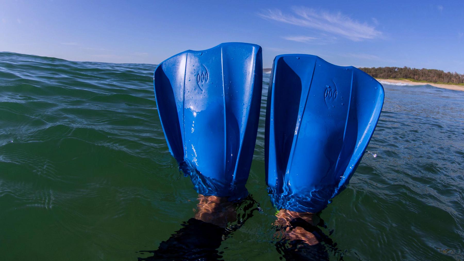 A rider's feet wearing blue POD PF2 swim fins raised above the water surface as seen through POD AquaLuxe silver mirror goggle lenses, with a sandy beach and tree line visible in the background under a clear blue sky