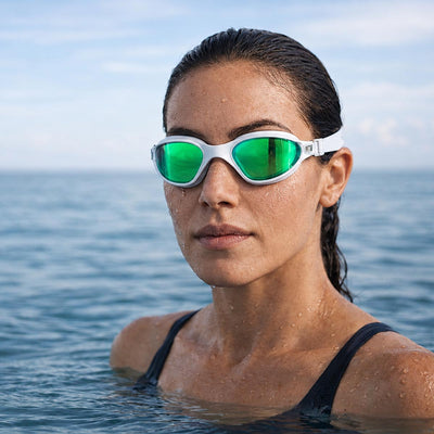 A female swimmer in open water wearing POD AquaLuxe swim goggles with green mirror lenses and a white frame, with water droplets on her face and open ocean in the background