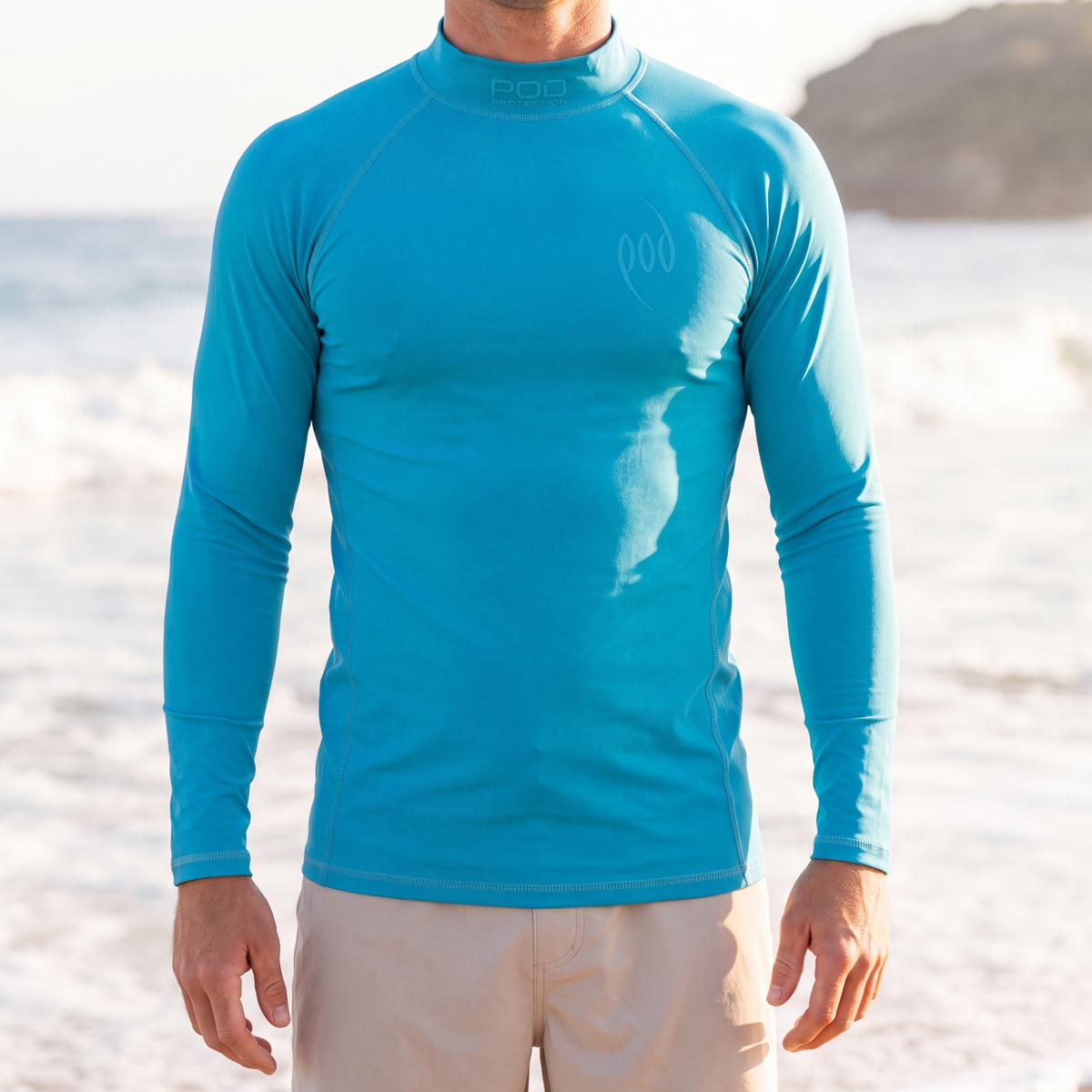 A man wearing a bright turquoise blue long-sleeve rash guard on a beach, with the ocean and shoreline blurred in the background.