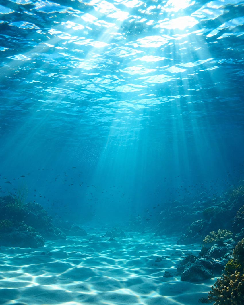 Underwater ocean scene with bright sunbeams filtering through the water surface and casting light on the seabed, with coral and marine life faintly visible in the blue water.