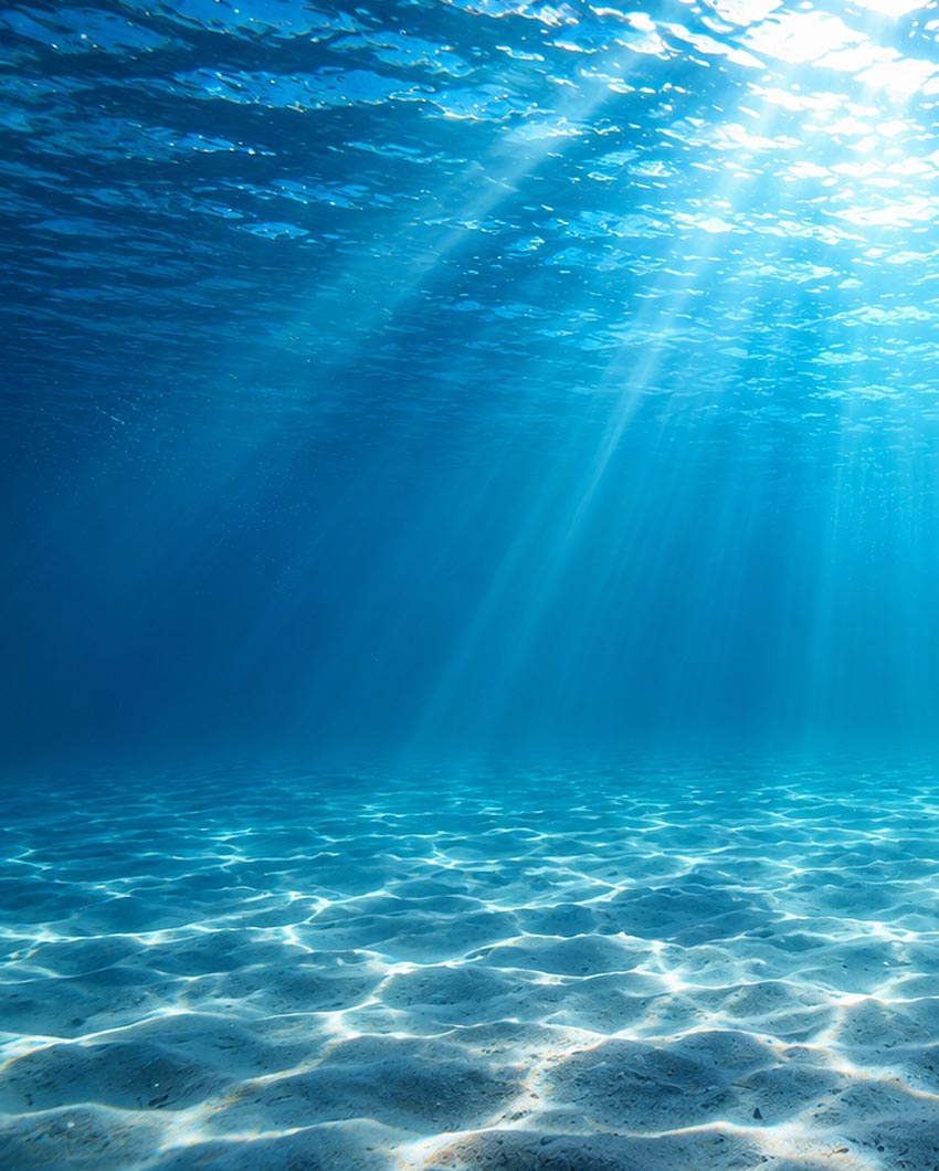 Underwater ocean scene with bright blue water, sun rays filtering through the surface, and sunlight rippling across the sandy seabed.