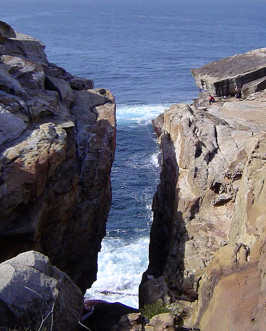 A scenic coastal view showing a narrow rocky gorge with steep tan and brown cliffs framing a deep blue ocean with white surf crashing below.
