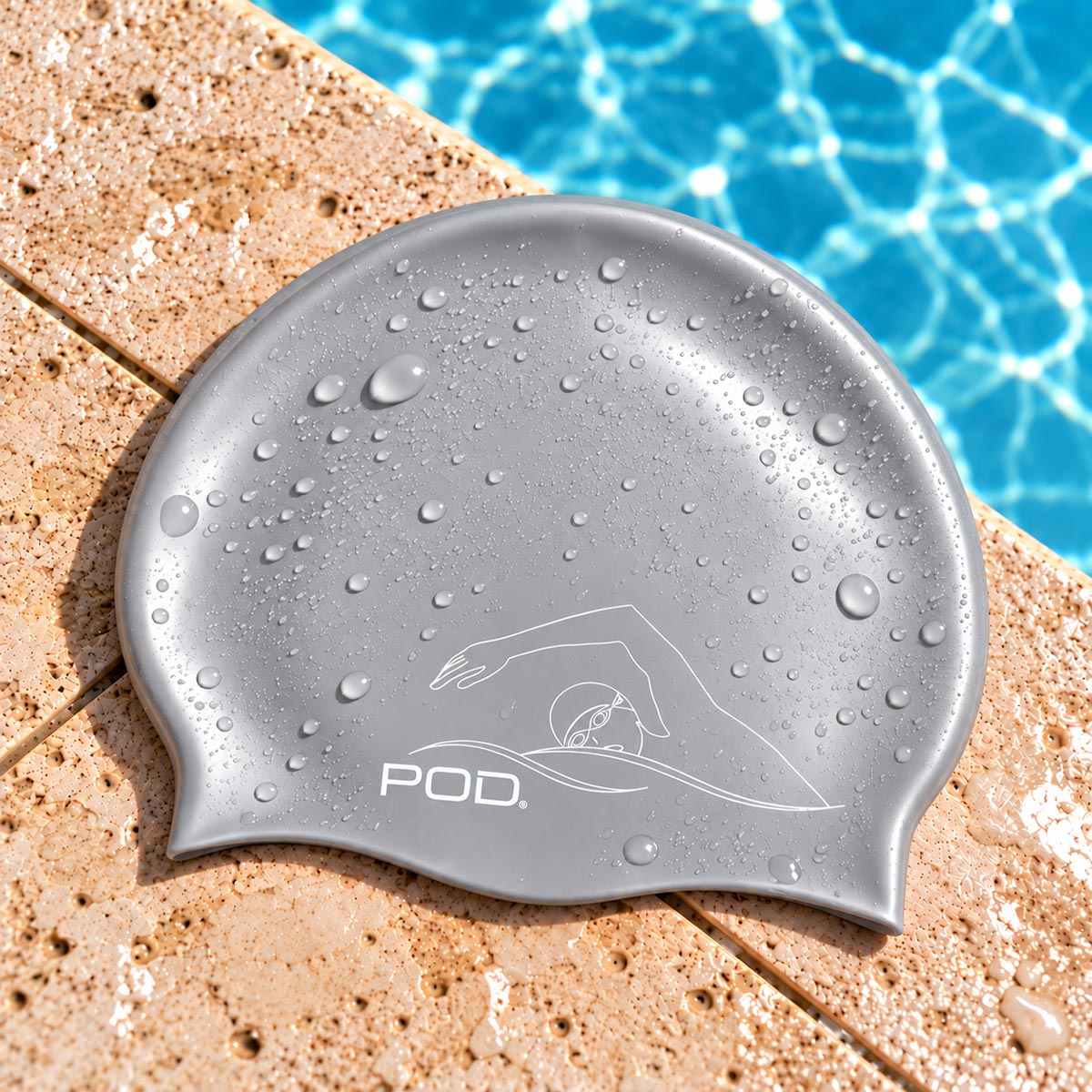 A gray silicone swim cap with water droplets, featuring a white line art logo and the brand name 'POD.' resting on a beige stone pool deck with blue pool water in the background.