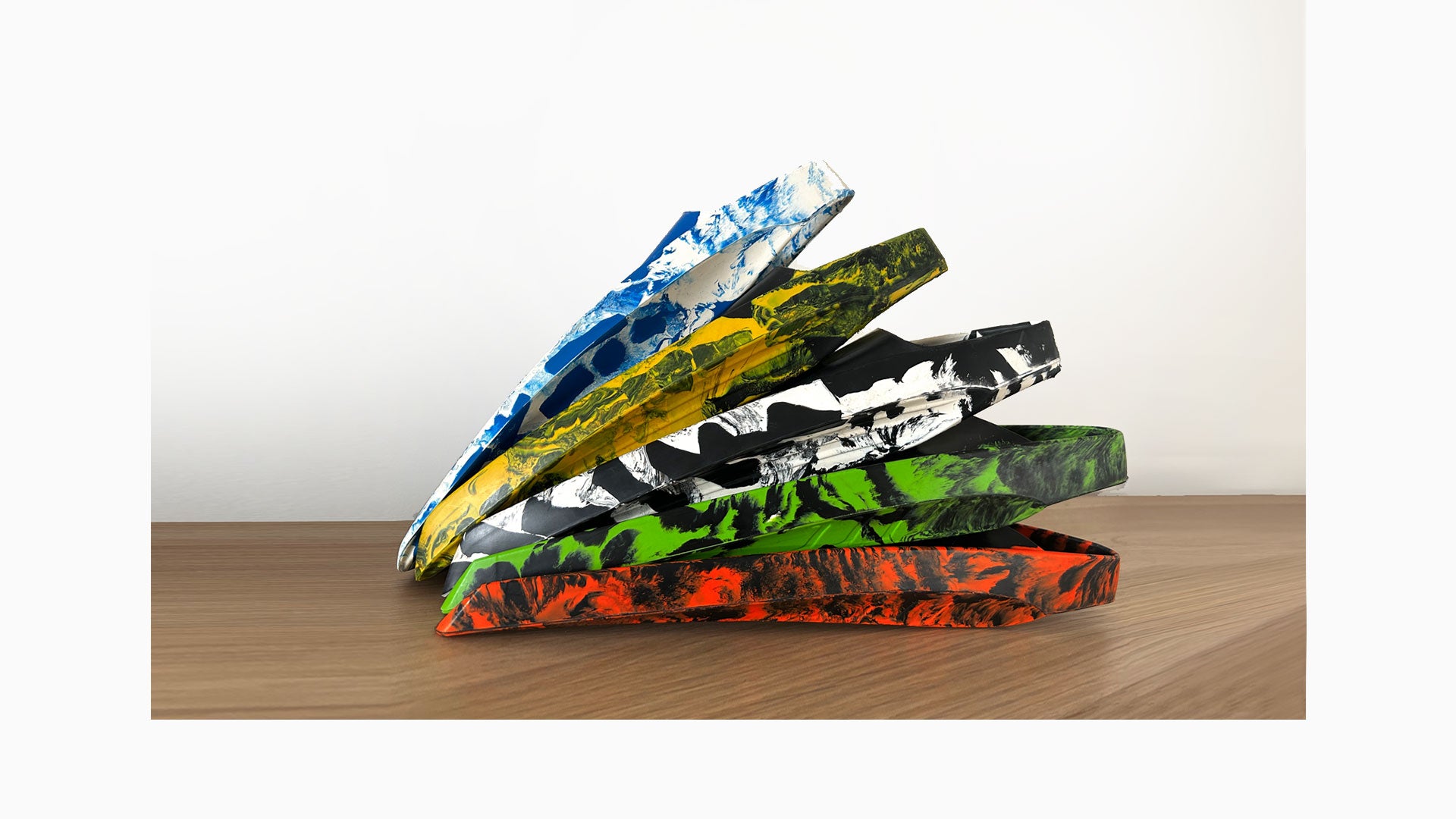 Five POD swim fins in variegated colour ways fanned and stacked on a timber surface, showing blue and white, yellow and black, black and white, green and black, and red and black natural rubber