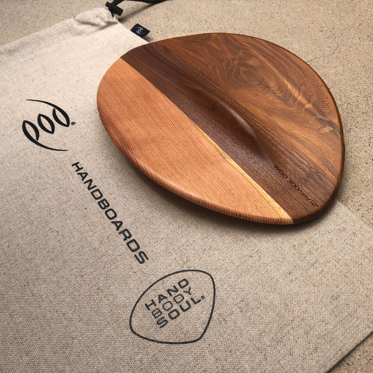 The WOO 300mm 12 inch cedar and walnut handboard resting on a canvas POD Handboards drawstring bag printed with the POD logo and Hand Body Soul mark
