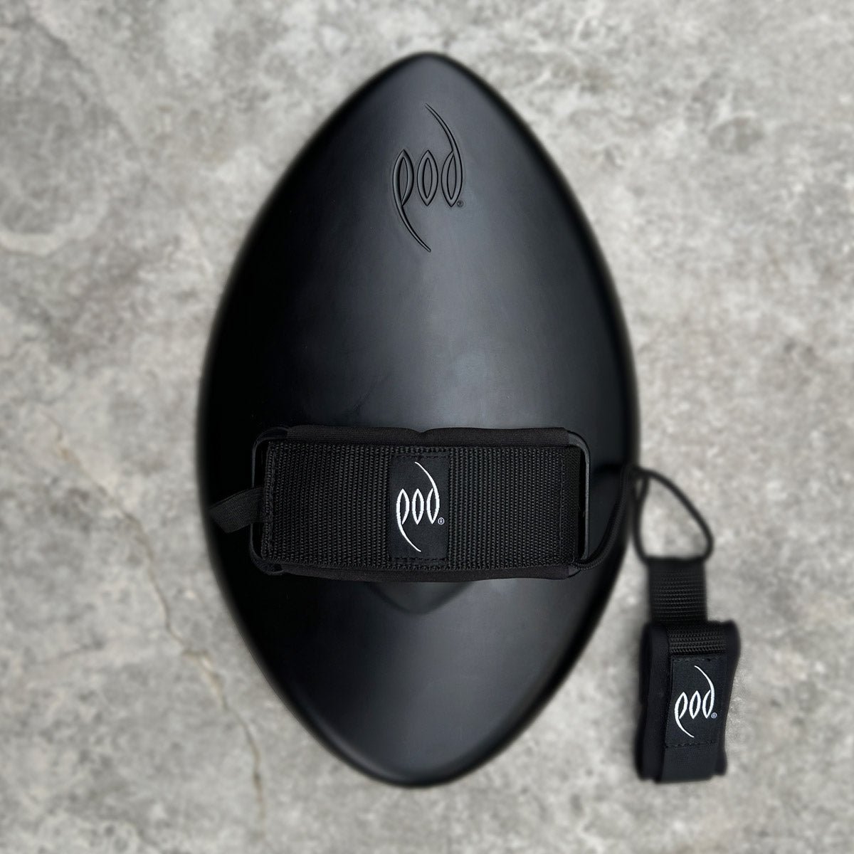 A black Pod brand handboard  bodysurfing handplane with an integrated wrist strap and matching ankle strap, shown on a textured gray stone background.