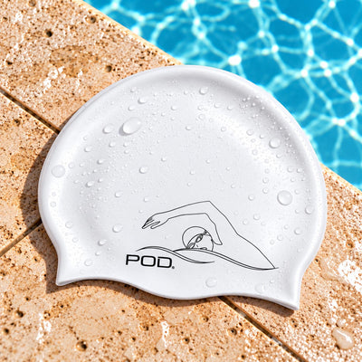 A white silicone swim cap with a black line-art logo and the text 'POD' on the front, covered in water droplets and placed on a pool deck with blue pool water in the background.
