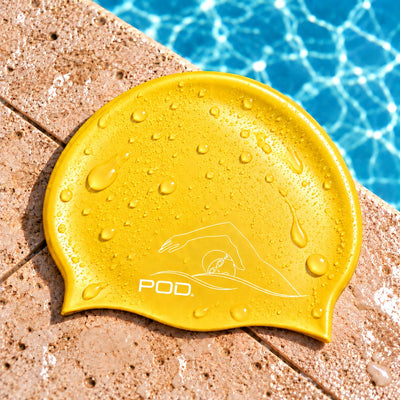 A yellow swim cap with a white POD logo and swimmer graphic, covered in water droplets, resting on a pool deck with blue pool water in the background.